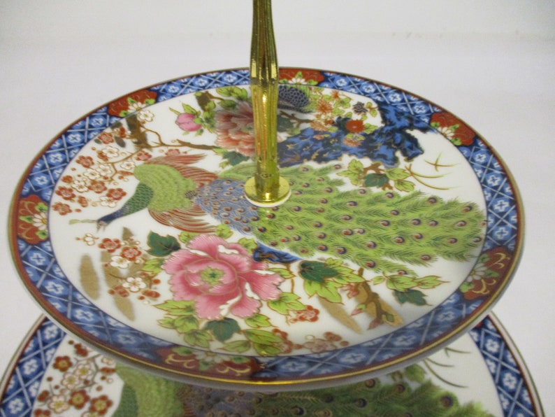 May include: Two-tiered porcelain serving stand with a gold metal handle. The stand features a white background with a colorful peacock design and floral accents.