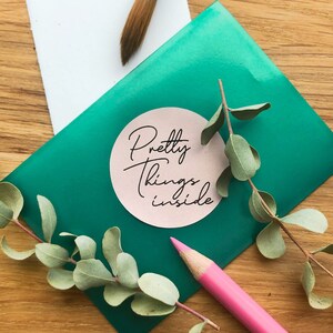 May include: A green envelope with a pink circle sticker that says "Pretty things inside". The envelope is on a wooden surface with a sprig of green leaves and a pink pencil.