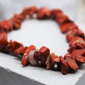 May include: A red gemstone bracelet made of irregular shaped stones. The bracelet is on a gray surface.