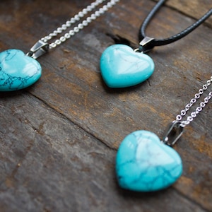 May include: Three turquoise heart-shaped pendants on silver and black chains. The pendants are on a wooden surface.