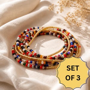 May include: A set of three beaded bracelets featuring a mix of small, multicolored glass beads in shades of red, blue, black, white, and yellow, interspersed with metallic gold cylindrical beads arranged in a circular pattern with a yellow starburst label reading SET OF 3.