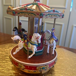 May include: A decorative carousel music box featuring a red and gold base, a canopy with green and red accents, and multiple white horses with riders. The central column is mirrored, and the figures are painted in blue, gold, and white tones.