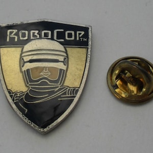 Op de afbeelding: Een gouden en zwarte emaille pin met het hoofd van Robocop en de tekst "Robocop" met een handelsmerk. De pin heeft een gouden metalen achterkant met een veiligheidsspeld.