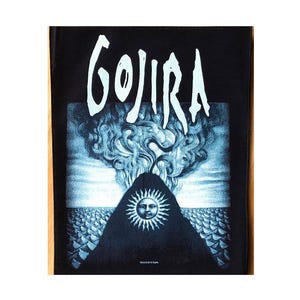 May include: Black rectangular patch featuring the band name "GOJIRA" in white, stylized lettering. Below, a detailed blue and white illustration depicts a mountain with a sun face, surrounded by swirling clouds and a textured sea.