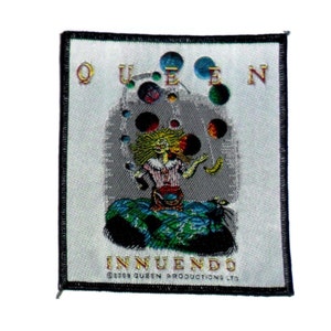 May include: A white fabric patch with black edging featuring a colourful illustration of a figure juggling balls. The text "QUEEN" is above the figure and "INNUENDO" is below the figure. The text "©1991 QUEEN PRODUCTIONS LTD" is at the bottom of the patch.