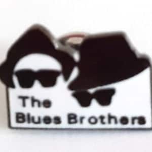 May include: A black and white enamel pin featuring a silhouette of two men wearing hats and sunglasses. The text "The Blues Brothers" is printed below the silhouettes.