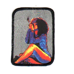 May include: A colourful embroidered patch featuring a woman with long hair sitting with her legs crossed. The background is a swirling black and white pattern.