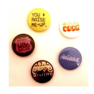 May include: Five round buttons with different designs. The first button is yellow with black text that says "You raise me up." The second button is white with red text that says "Westlife Club." The third button is brown with a pink design that says "WB6." The fourth button is black with a white design of four faces and the text "Westlife." The fifth button is blue with white text that says "Westlife."