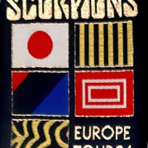 May include: A black patch with the word "SCORPIONS" in gold lettering. Below the word are four squares with different designs: a white square with a red circle, a yellow and black striped square, a blue and black square with a red triangle, and a red square with a white square inside. Below the squares is the text "EUROPE TOUR 84" in gold lettering.