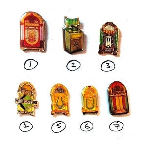 Könnte beinhalten: Eine Sammlung von sieben Emaille-Anstecknadeln in Form von Jukeboxen. Die Anstecknadeln weisen verschiedene Farben auf, darunter Rot, Gold, Grün und Gelb. Einige Anstecknadeln tragen den Text "POLEPOSITION". Die Anstecknadeln sind auf einer weißen Oberfläche angeordnet.
