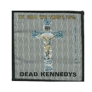 May include: A black and white embroidered patch with a grey background. The patch features a cross with a figure of Jesus Christ on it. The text "In God We Trust, Inc." is at the top of the patch and "Dead Kennedys" is at the bottom.