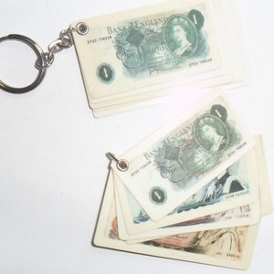 Könnte beinhalten: Ein Schlüsselanhänger mit einem Stapel Miniatur-Nachbildungen britischer Ein-Pfund-Noten mit einem Porträt von Königin Elisabeth II. Die Noten sind mit dem Text "Bank of England" und der Seriennummer "E122 759228" bedruckt.