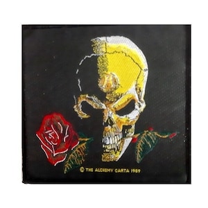 Peut inclure: Écusson brodé avec un crâne jaune et une rose rouge sur fond noir. Le texte "© THE ALCHEMY CARTA 1989" est visible en bas. Parfait pour personnaliser des vestes, sacs ou autres textiles. Dimensions adaptées pour une application facile.