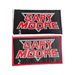 May include: Two rectangular patches with the text "GARY MOORE" in red, outlined in white, on a black background. The patches have a red border. The number "1" is on the top patch, and "2" is on the bottom patch.
