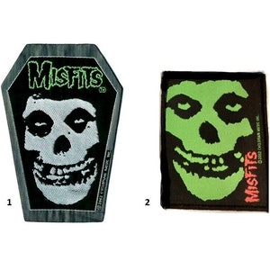 May include: Two Misfits patches. One is coffin-shaped with a white skull and the word "Misfits" in green. The other is rectangular with a green skull and the word "Misfits" in red. Both have the copyright information.