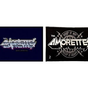 May include: Two black and white patches featuring the band name "The Amorettes" in a stylised font. The first patch shows the band name in a silver colour with a purple outline. The second patch has the band name in a white colour with a silver outline and the text "Keep it Hot" and "Keep it Heavy" in a smaller font.