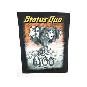 May include: Black patch featuring the band Status Quo. The design includes the band's name in yellow block letters above a tree-like structure with four faces. The background has an orange and grey gradient.