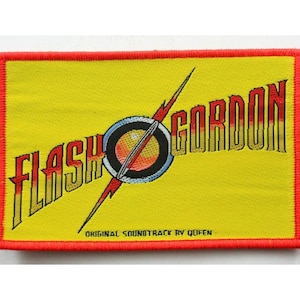 May include: A yellow embroidered patch with a red border. The patch features the text "FLASH GORDON" in a stylised font with a red and yellow lightning bolt and a red, yellow, and blue planet. The text "ORIGINAL SOUNDTRACK BY QUEEN" is printed in black below the graphic.
