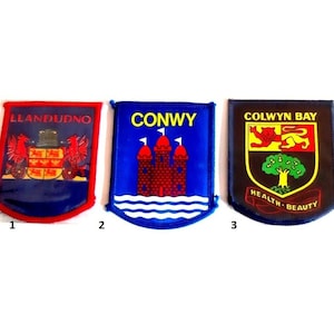 May include: Three embroidered patches with different designs. The first patch is red and blue with the text "LLANDUDNO" and a coat of arms. The second patch is blue with the text "CONWY" and a castle. The third patch is brown and blue with the text "COLWYN BAY" and a coat of arms with a tree.
