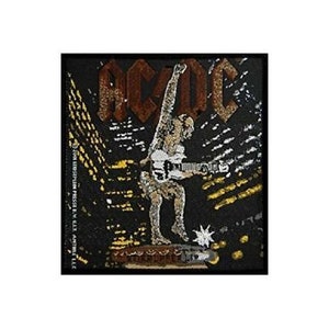 May include: Black and white embroidered patch featuring the word "AC/DC" in brown lettering. A figure in a white shirt and black trousers is playing a guitar against a yellow and black background.