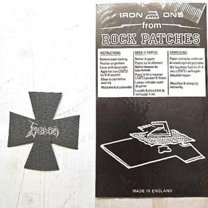 May include: A black iron-on patch in the shape of a Maltese cross with the word "Venom" in silver lettering. The patch is next to a black and white package with instructions for application and the text "IRON ONS from ROCK PATCHES".