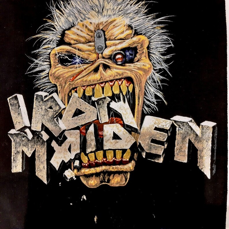 May include: A black t-shirt featuring a graphic of a metal band mascot. The mascot is a monstrous, grey-haired figure with a large mouth and a mechanical eye. The band's name, "IRON MAIDEN", is written in large, silver letters across the front of the shirt.