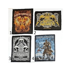 May include: Four rectangular embroidered patches featuring Amon Amarth designs. Patch 1 shows orange wings and text. Patch 2 displays a yellow skull and text. Patch 3 features a white hammer and skulls. Patch 4 depicts a warrior with an axe in blue tones.