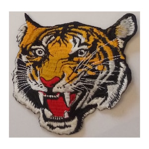 May include: A close-up of an embroidered tiger head patch. The tiger has yellow and orange striped fur with black stripes and a red mouth.