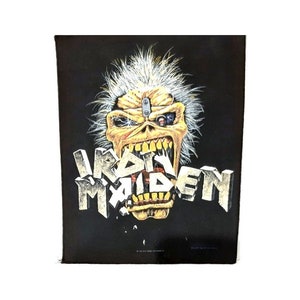 May include: Black patch featuring a yellow and white illustration of a monster with a metal plate on its forehead and a mechanical eye. The monster's mouth is open and the text "IRON MAIDEN" is in front of it.