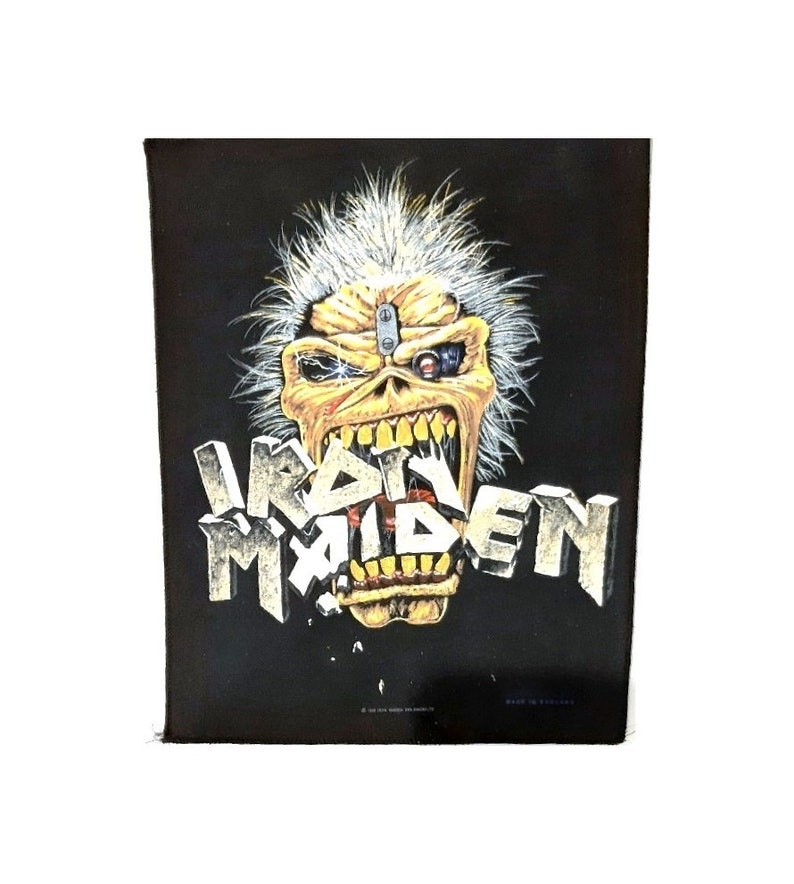May include: Black patch featuring a yellow and white illustration of a monster with a metal plate on its forehead and a mechanical eye. The monster's mouth is open and the text "IRON MAIDEN" is in front of it.