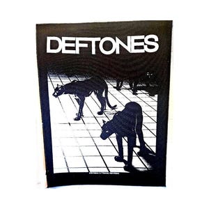 May include: Black and white patch featuring the band name "DEFTONES" in bold white letters. The patch design includes black panther silhouettes on a white grid background. The patch is rectangular.