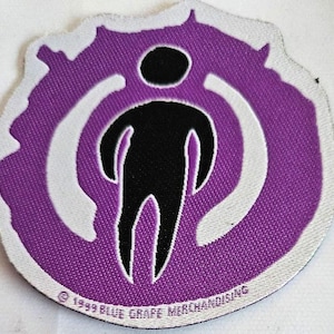 May include: Purple and white embroidered patch with a stylized black figure in the center. The figure is standing inside a circle with a white outline. The text "1999 BLUE GRAPE MERCHANDISING" is at the bottom of the patch.