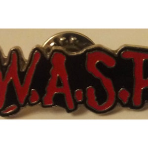May include: A black and red metal pin with the word "W.A.S.P." in a stylized font.