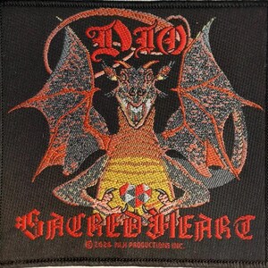 May include: A black embroidered patch with a red, orange, and yellow dragon with wings spread wide. The dragon is holding a red, white, and black symbol in its claws. The text "DIO" is above the dragon and "SACRED HEART" is below the dragon. The text "© 1983 M.I.H. PRODUCTIONS INC." is at the bottom of the patch.