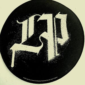 May include: Circular black patch with a white 'LP' design in a gothic font. The design has a distressed, spray-painted effect. The patch is likely a music-related item, possibly a band logo or merchandise.