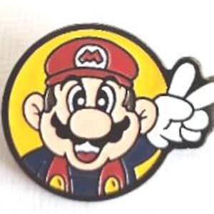 May include: A round enamel pin featuring a cartoon image of Mario from the Super Mario Bros. video game franchise. Mario is wearing his signature red cap and overalls, with a white-gloved hand making a peace sign. The pin has a yellow background.