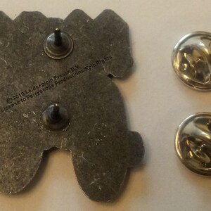 May include: A silver metal pin featuring a cartoon image of a video game controller. The back of the pin has the text "&copy;2019 Leidscheplein Press B.V. Under License to Periscope Productions/Epic Rights".