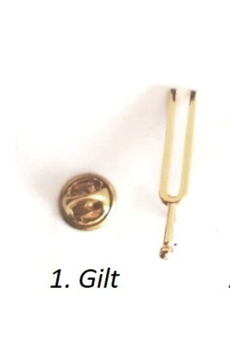 Puede incluir: Un diapas&oacute;n dorado y un alfiler dorado con cabeza redonda. El texto "1. Gilt" es visible debajo del alfiler.