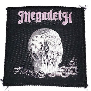 May include: Black and white embroidered patch with the band name "Megadeth" in pink lettering. The patch features a skull with a detailed design.