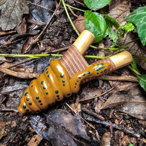 May include: A handcrafted wooden slingshot with a coiled snake design. The snake is painted in shades of yellow and brown with blue spots. The slingshot's frame is made of natural wood, and the handle is wrapped with brown twine.
