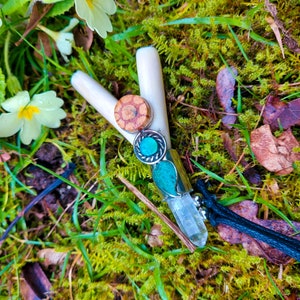 May include: A handcrafted wand with a clear quartz crystal, turquoise, and wooden accents. The wand is set against a backdrop of green moss and wildflowers, with a black cord attached. The wand's design incorporates natural elements and earthy tones.