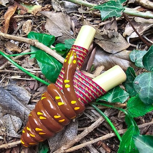 May include: A handmade slingshot with a brown, snake-like grip and yellow spots. The wooden arms are wrapped with red, green, and yellow twine. The slingshot is set against a backdrop of leaves and twigs.