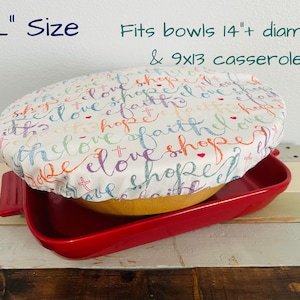 Reusable bowl cover in "Faith Hope Love" print. An XL size, white bowl cover with colorful script that reads "love, hope, faith". The cover is placed over a yellow bowl.  The bowl is placed on top of a red casserole dish. Text in image reads: "Fits bowls 14"+ diameter & 9x13 casseroles".