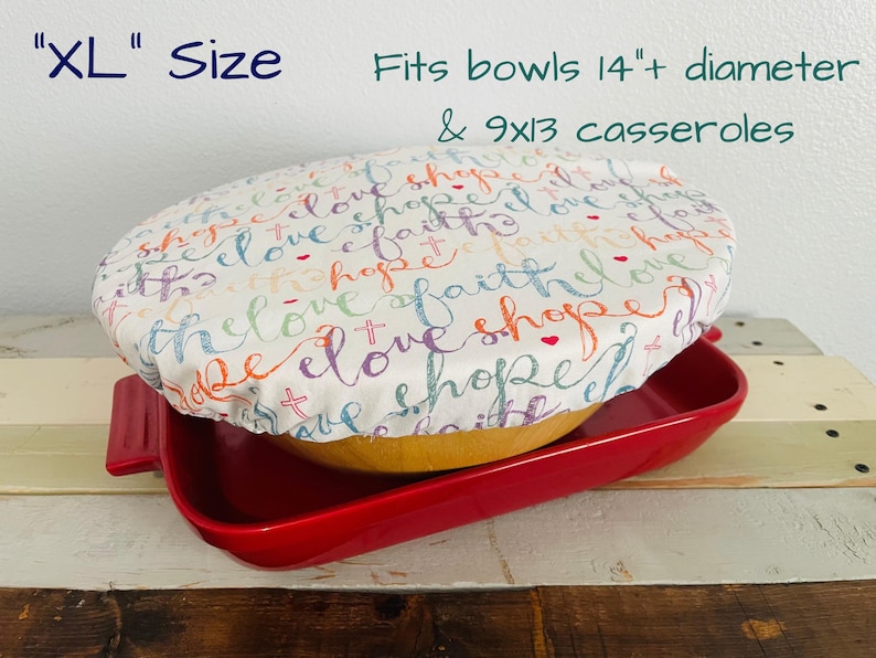 Reusable bowl cover in "Faith Hope Love" print. An XL size, white bowl cover with colorful script that reads "love, hope, faith". The cover is placed over a yellow bowl.  The bowl is placed on top of a red casserole dish. Text in image reads: "Fits bowls 14"+ diameter & 9x13 casseroles".