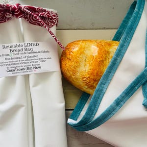 May include: White reusable bread bag with a red and white patterned tie closure. The bag has a label that reads "Reusable LINED Bread Bag". A loaf of bread is visible inside a white bag with teal straps.