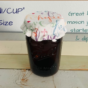 A glass mason jar with a fabric lid featuring "Faith Hope Love" print - a colorful script and a cross design. The jar is labeled "MINI/CUP" and "Size". The text "Great for mason jars, starters, & dips" is also visible.