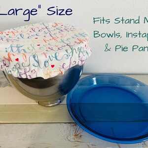 Reusable "Large" sized fabric bowl cover in "Names of Jesus" print: white fabric with the words "love", "hope", and "faith" in various colors.  The cover is on a stainless steel stand mixer bowl. A blue pie pan sits next to the bowl. Text on the image reads "Large" Size and "Fits Stand Mixer Bowls, Instapots, & Pie Pans."