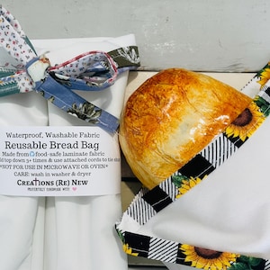May include: Two white reusable bread bags made of waterproof fabric. One bag is tied with a floral patterned cord and features a label reading Waterproof, Washable Fabric Reusable Bread Bag. The other bag has a sunflower and plaid trim.