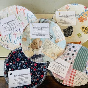 May include: Five circular fabric jar grip openers with various patterns including floral, chicken, kitchen utensil, and script prints. Each item features a white tag labeled Jar Grip Openers with text describing their use for opening jars.