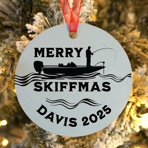 May include: A round, white Christmas ornament with a red ribbon. The ornament features a black silhouette of a fishing boat with a person fishing, the words "MERRY SKIFFMAS DAVIS 2025" in black, and a wavy water design.